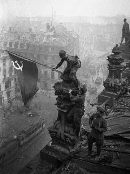 Fotografia in bianco e nero: soldati sovietici issano una bandiera con falce e martello sulla sommità del Reichstag in macerie, con una strada e un tram sullo sfondo